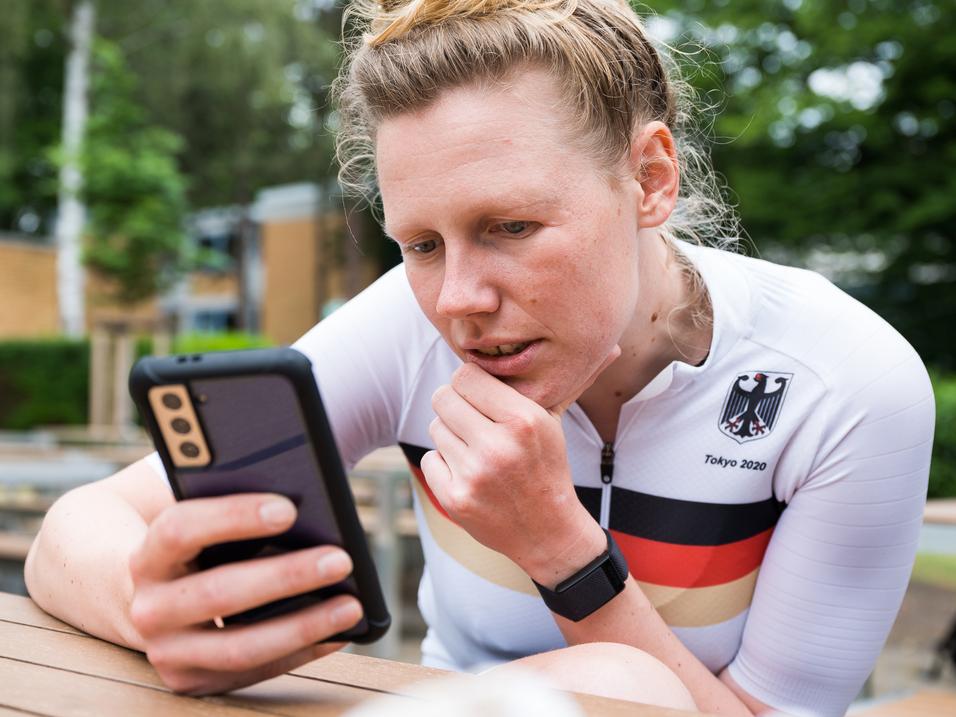 Athletin im weißen Trikot mit schwarzem und rotem Streifen schaut konzentriert auf ihr Smartphone in einer Außenanlage.